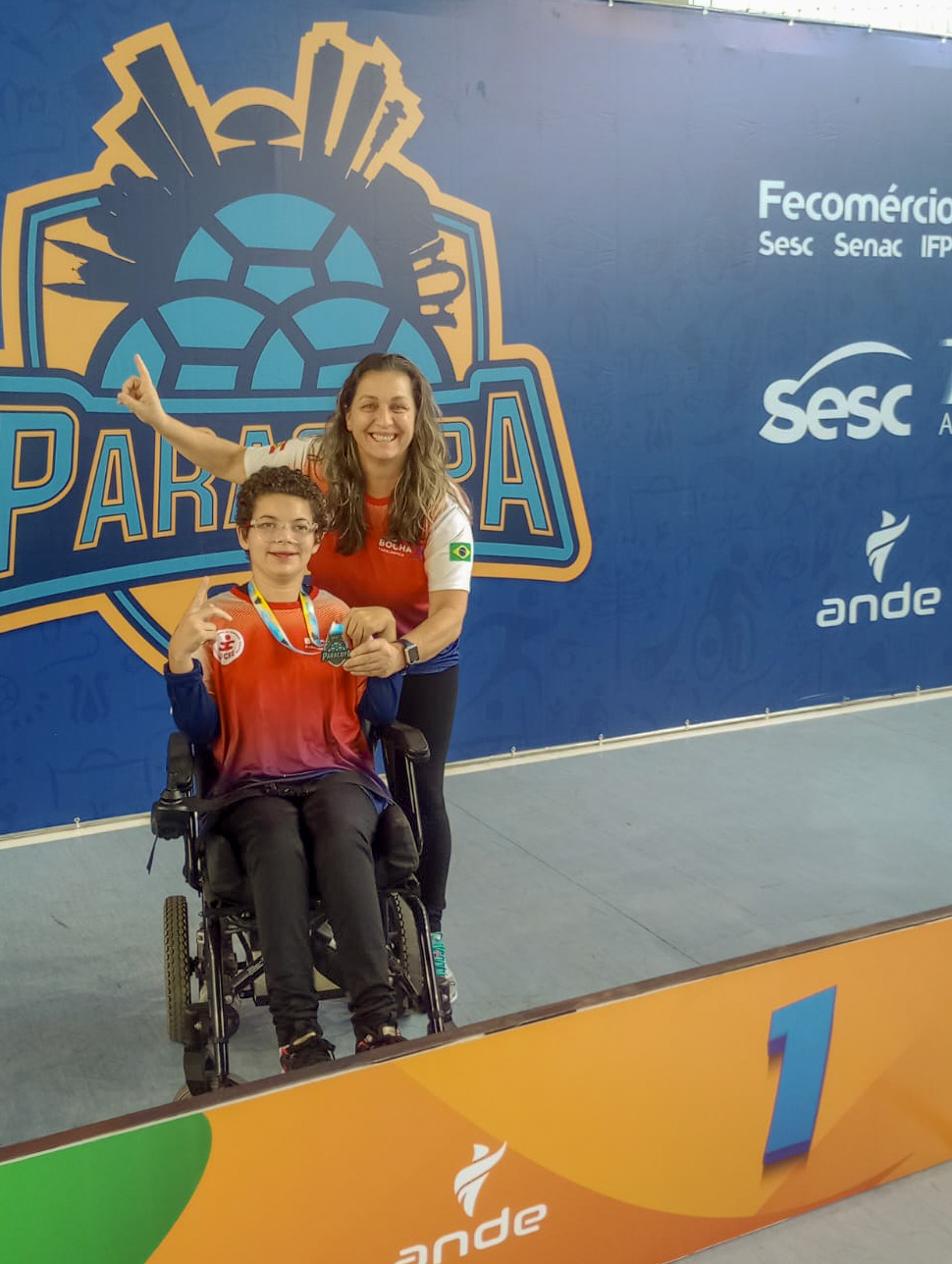 duas pessoas, uma sentada em cadeira de rodas e outra em pé, com as mãos pra cima segurando medalha