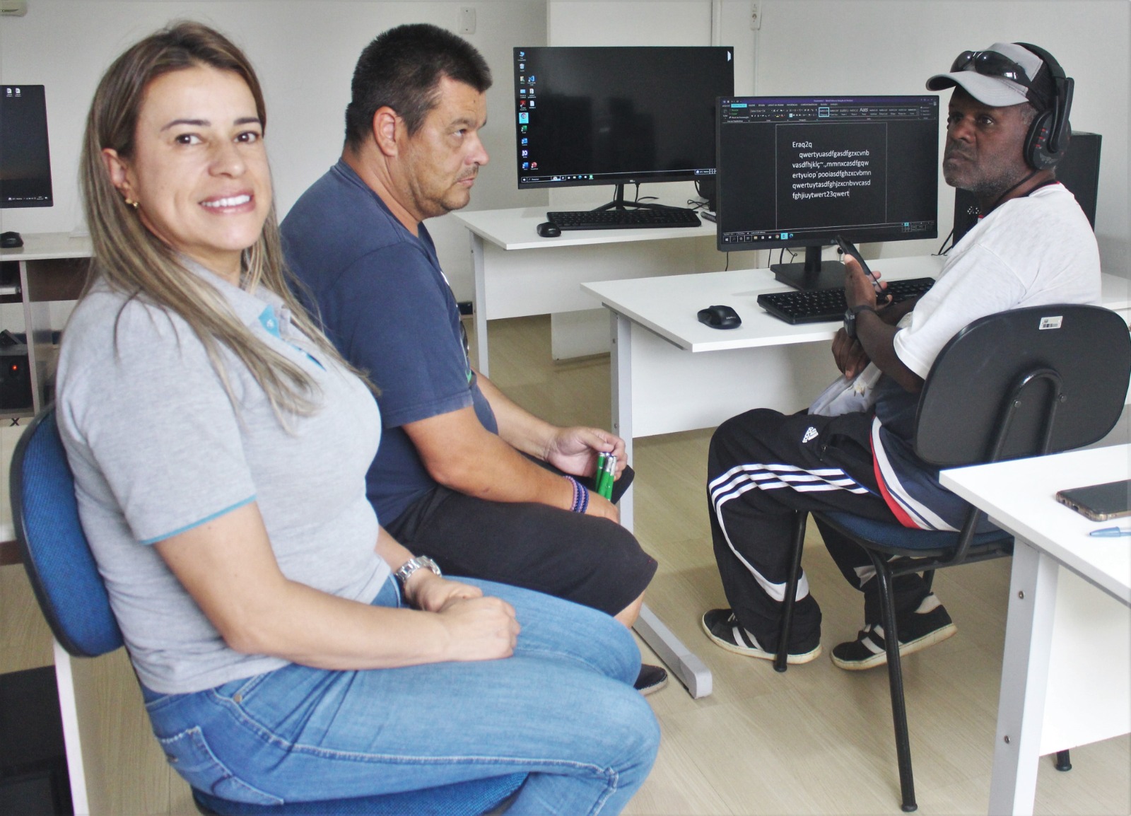 Ambiente interno, 3 pessoas sentadas, mesa com computador e fones de ouvido. 