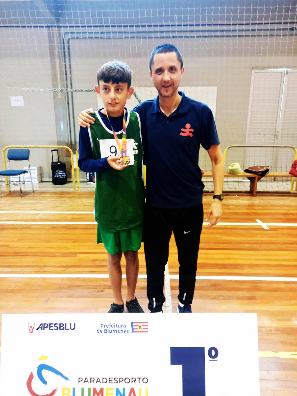 Um menino mostrando medalha e um homem em pé posam para foto abraçados, dentro de ginásio de esportes. 