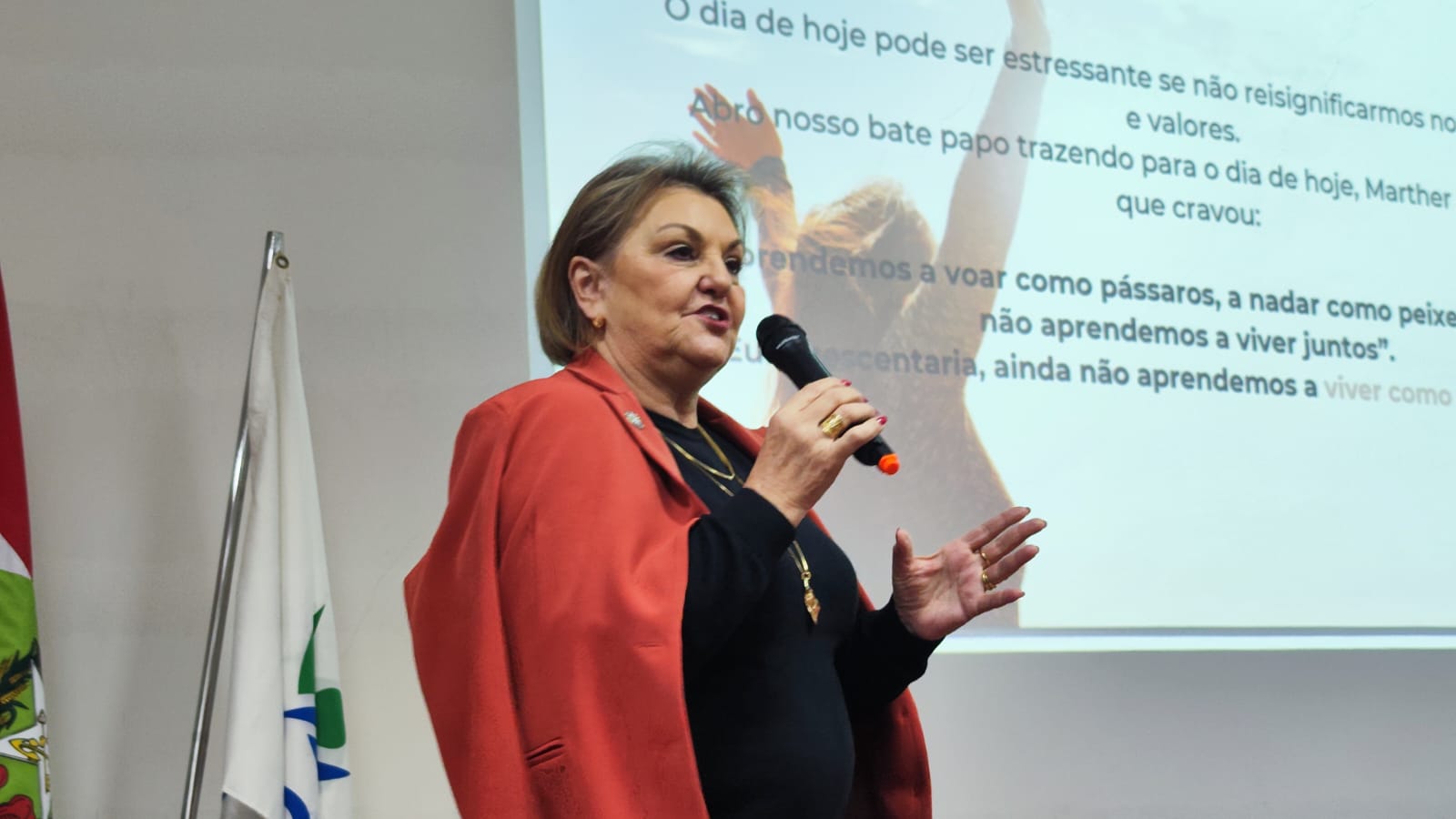 Mulher falando ao microfone com telão ao fundo 