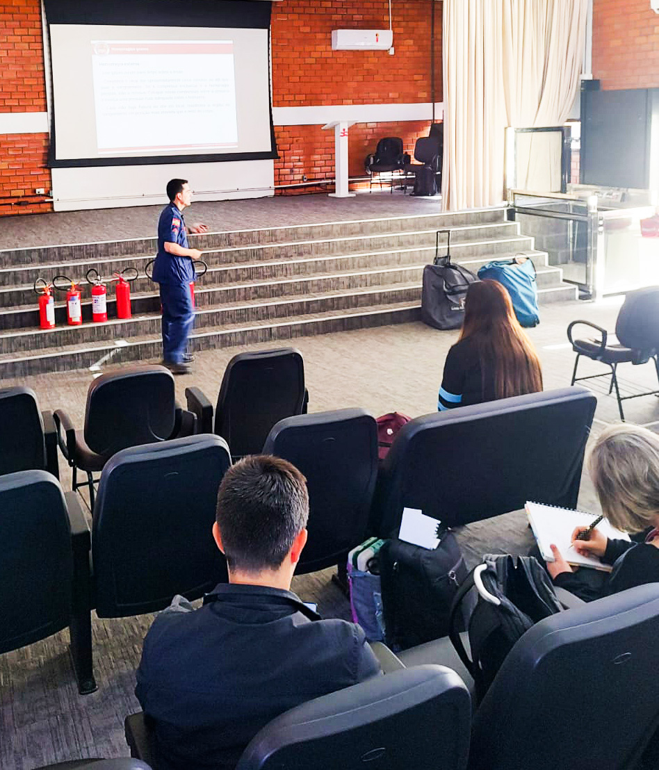 Imagem de auditório, um homem com farda azul escuro, ao fundo 4 extintores no chão. pessoas na plateia assistindo. 