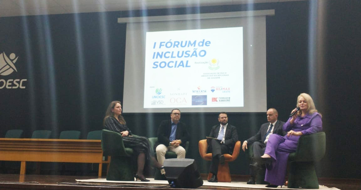 Cinco pessoas sentadas, palco de auditório, falando ao microfone