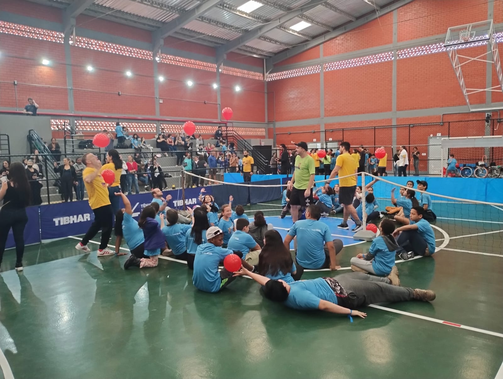 Um grupo de pessoas estão sentadas no centro de uma quadra esportiva,  de cor verde. Uma pessoa, que aparece em primeiro plano está deitada. Todos usam camisetas azuis. Ao fundo aparece três pessoas em pé com camisetas amarelas próximo a uma rede de badminton